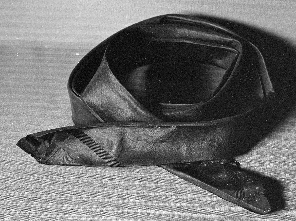 Svart, litt stivt silketørkle brettet som halstørkle. Full vevbredde. Faldet inn for hånd på to kanter.