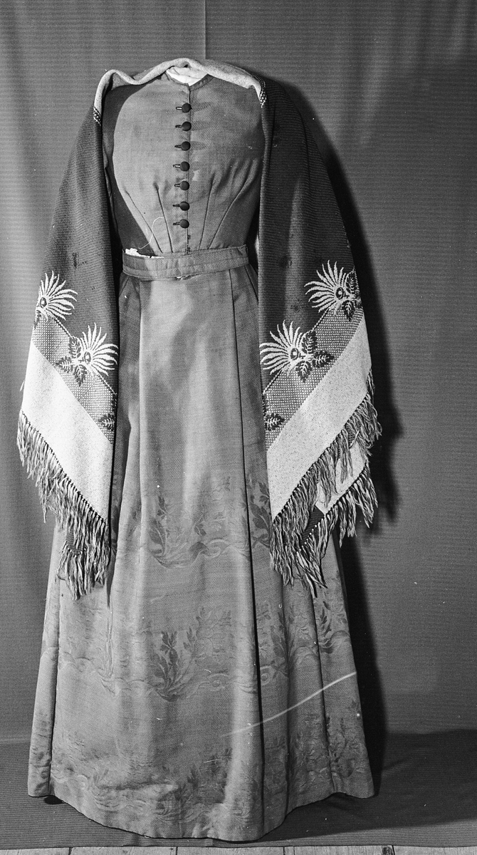 Todelt kjole i tynn brokade med blomstermønster innvevd på skjørtet. Kjolelivet er helfòret med to ulike stoffer. ermene er sydd i for hånd og sårkantene kastet over for hånd. stikningssømmer er sydd på maskin. Sidesømmer og innsnitt med lett spiling likeså. Kjolelivet lukkes foran med stofftrukne knapper og to hekter nederst. Nederst på ermene er det pyntet med oppbrett med grønne bånd, kapper og en effekt i kjolens stoff. Ermene er skåret i to deler.
Skjørtet er foldet foran med splitt i fold på venstre side som lukkes med hekter på to steder. Nederst på innsiden er det skoningskant og slitesnor. Bak i livet er det et felt på 6 cm med striping.
Belte med hektelukning under sløyfe som har sittet bak.
Kjøpt stoff. Muligens brukt som modell for stakk til Sør-Østerdalsbunaden.