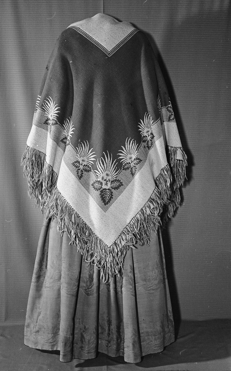 Todelt kjole i tynn brokade med blomstermønster innvevd på skjørtet. Kjolelivet er helfòret med to ulike stoffer. ermene er sydd i for hånd og sårkantene kastet over for hånd. stikningssømmer er sydd på maskin. Sidesømmer og innsnitt med lett spiling likeså. Kjolelivet lukkes foran med stofftrukne knapper og to hekter nederst. Nederst på ermene er det pyntet med oppbrett med grønne bånd, kapper og en effekt i kjolens stoff. Ermene er skåret i to deler.
Skjørtet er foldet foran med splitt i fold på venstre side som lukkes med hekter på to steder. Nederst på innsiden er det skoningskant og slitesnor. Bak i livet er det et felt på 6 cm med striping.
Belte med hektelukning under sløyfe som har sittet bak.
Kjøpt stoff. Muligens brukt som modell for stakk til Sør-Østerdalsbunaden.