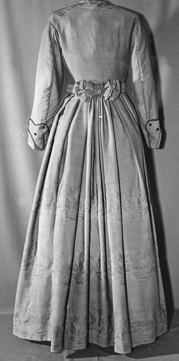 Todelt kjole i tynn brokade med blomstermønster innvevd på skjørtet. Kjolelivet er helfòret med to ulike stoffer. ermene er sydd i for hånd og sårkantene kastet over for hånd. stikningssømmer er sydd på maskin. Sidesømmer og innsnitt med lett spiling likeså. Kjolelivet lukkes foran med stofftrukne knapper og to hekter nederst. Nederst på ermene er det pyntet med oppbrett med grønne bånd, kapper og en effekt i kjolens stoff. Ermene er skåret i to deler.
Skjørtet er foldet foran med splitt i fold på venstre side som lukkes med hekter på to steder. Nederst på innsiden er det skoningskant og slitesnor. Bak i livet er det et felt på 6 cm med striping.
Belte med hektelukning under sløyfe som har sittet bak.
Kjøpt stoff. Muligens brukt som modell for stakk til Sør-Østerdalsbunaden.