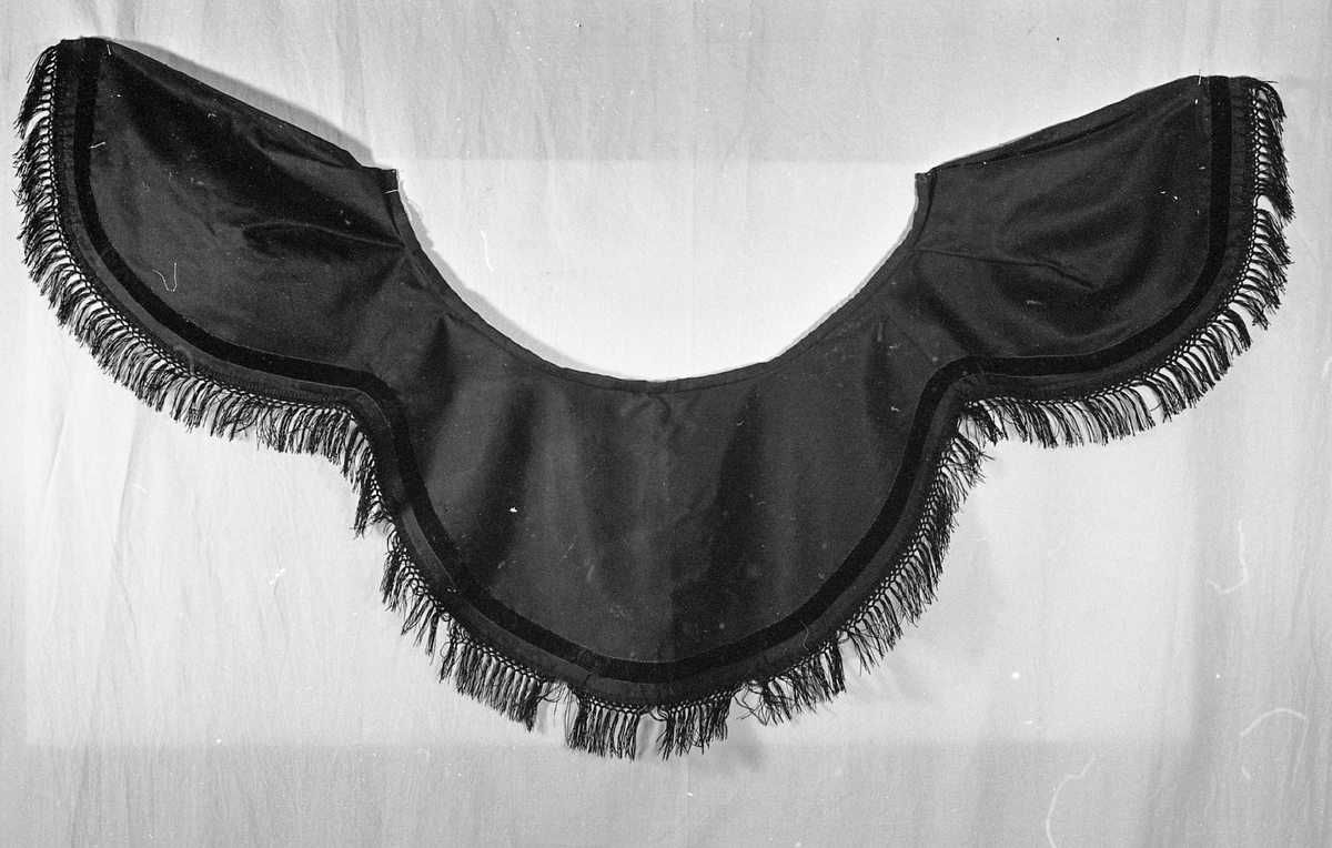 Krage som henger over skuldrene og er rund i formen. Den har fløyelsbånd og i nedkant bånd med frynser. Festes med hekter. 