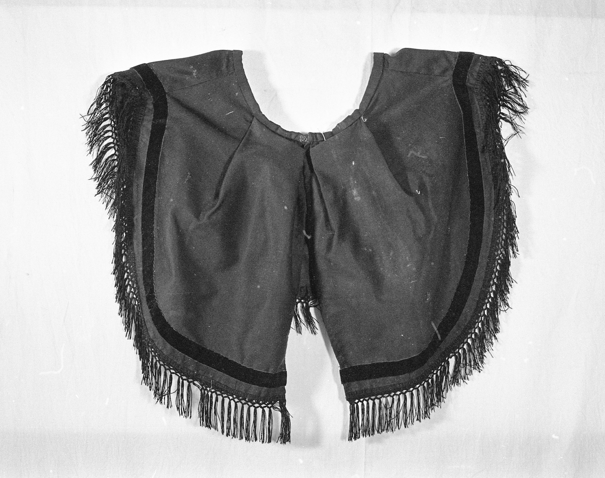 Krage som henger over skuldrene og er rund i formen. Den har fløyelsbånd og i nedkant bånd med frynser. Festes med hekter. 