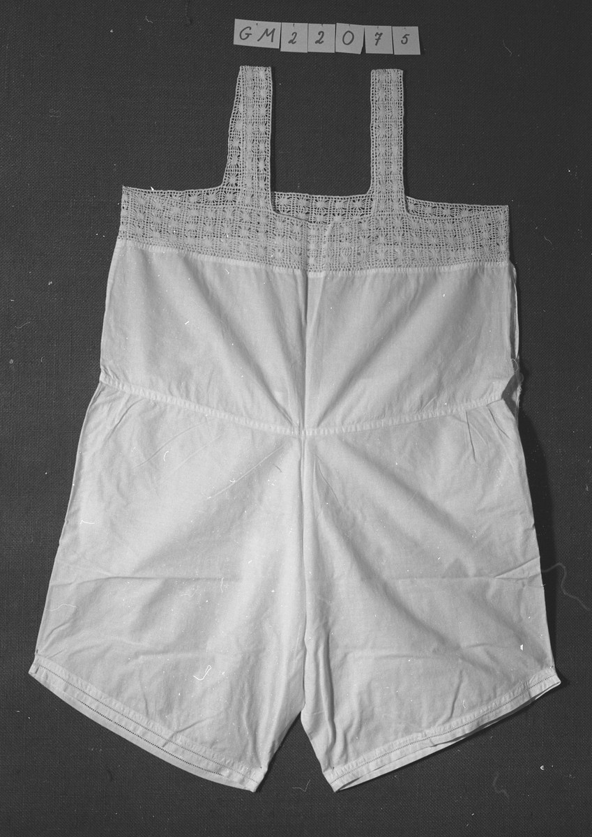 Combination, kvinne. N1b
Ett for-og ett bakstykke, rett oventil, påsydd 6 cm bred heklet kant rundt åpning. Ikke ermer, 3, 5 cm bred heklede skulderstropper. Buksa søm midt foran, 14 l splitt i sidene, kneppet med stofftrukken knapp, søm midt bak, bakstykket til å kneppe ned, 4, 5 cm bred linning, flere små rynkinger på hver side av midtsøm bak. Hullfaldsøm rundt låråpning. Livstykket sprukket i siden fra linning til heklekant.