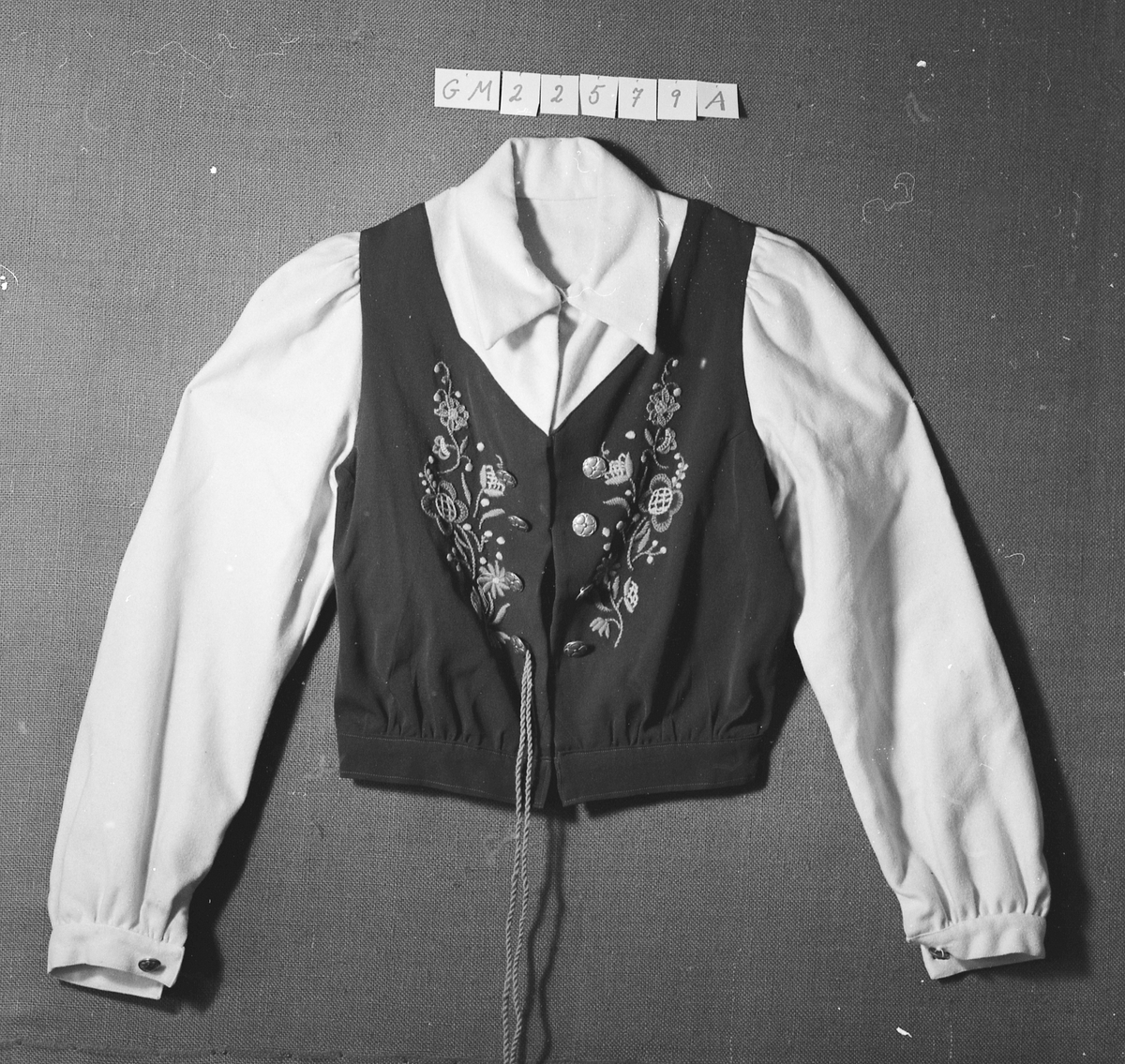 Overdel til drakt bestående av et kypertvevd blått bomullstoff brodert med ullgarn og med ermer og krage av lerretsvevd ull. Front og ermer har tinnknapper med støpt mønster. Det er hekter og trykknapper til å lukke med.
Buksa til drakta er av typen strekkbukse med press og strikk under beina. Den har glidelåslukning i siden og to lommer med glidelås i front.