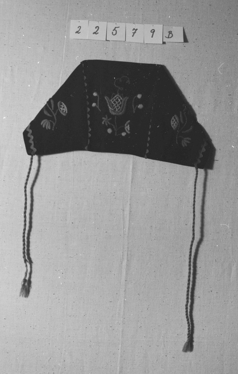 Overdel til drakt bestående av et kypertvevd blått bomullstoff brodert med ullgarn og med ermer og krage av lerretsvevd ull. Front og ermer har tinnknapper med støpt mønster. Det er hekter og trykknapper til å lukke med.
Buksa til drakta er av typen strekkbukse med press og strikk under beina. Den har glidelåslukning i siden og to lommer med glidelås i front.