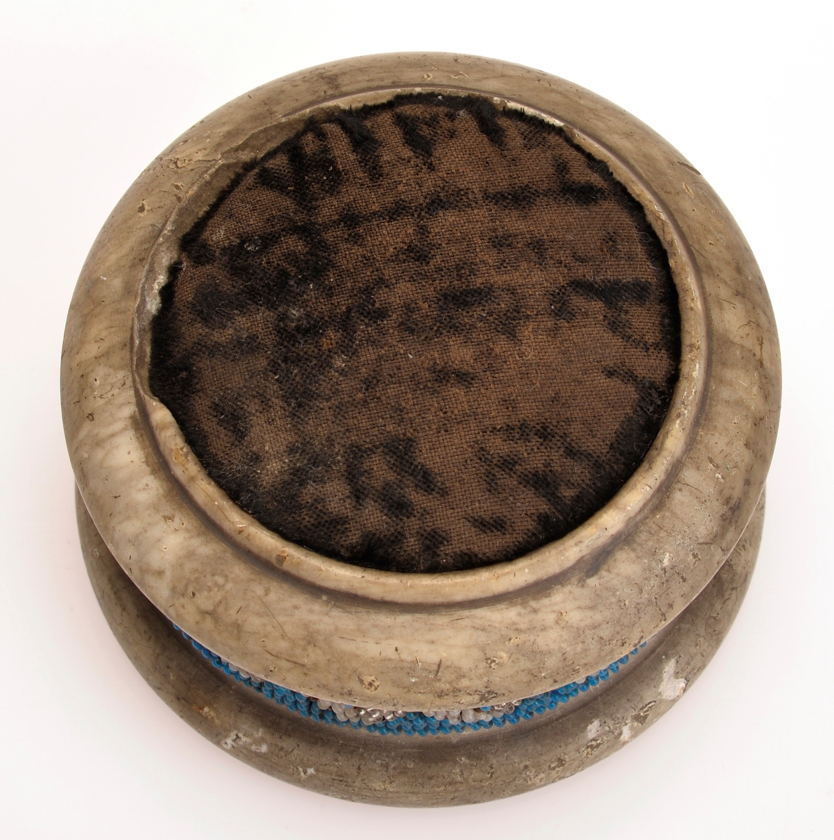 Rundformet kleberstein med nålepute av sort fløyel på toppen. Pyntet med mønstret perlebånd med hvite, blå og gjennomsiktige glassperler på midten. Perlene er sydd på stramei.