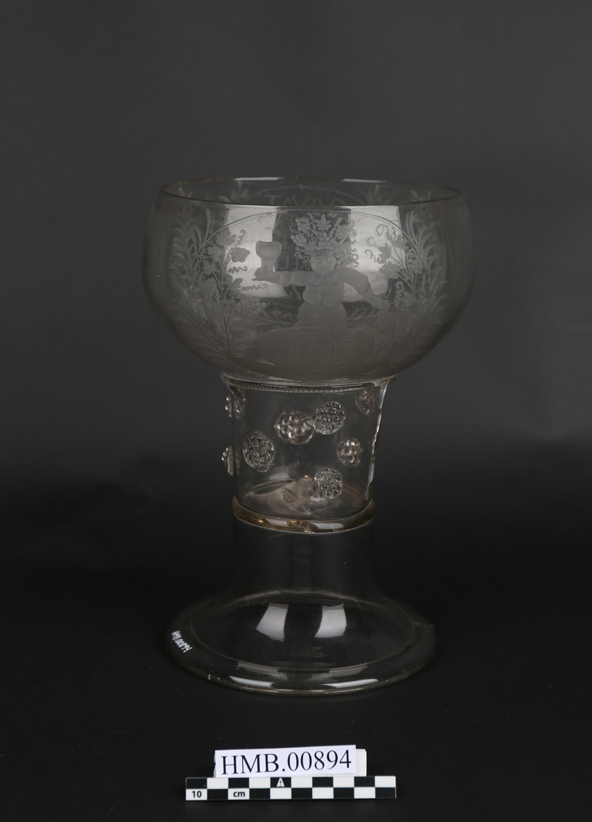 Pokal i glass. Sokkelen er to deler satt sammen oppå hverandre, med en bolle på toppen. Det er inngravert tekst og motiver rundt siden på bollen. Motivet er vinguden Bacchus som sitter på en tønne. Han har vindruer i den ene hånda og en pokal i den andre.