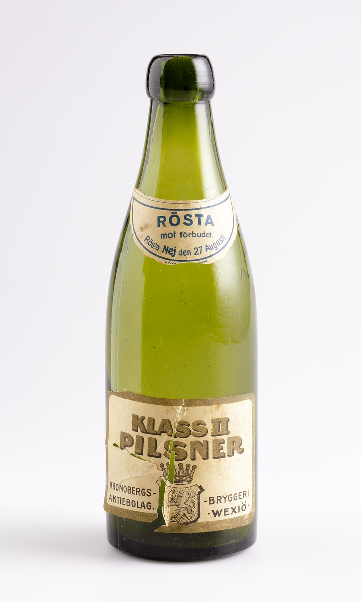 Ölflaska 1/3 liter. Tvådelad form med driven halsring. Relief på bröstet H 1/3 LIT. Månformad etikett med text; Rösta mot förbudet. Rösta Nej den 27 Augusti. Rektangulär etikett med text och Växjö stadsvapen med krona; Klass II Pilsner Kronobergs-Bryggeri Aktiebolag Växjö. Relief i botten,1921.

Inskrivet i huvudkatalogen efter 1984.