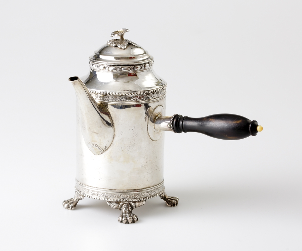 Kaffekanna i silver. 
Cylindrisk modell på tre tassfötter. Graverad bård nedtill, följd av pärlbård. Välvt lock med graverad bård, pärlbård, äggstavsbård och knopp i form av en ros. Försedd med svarvat, svärtat trähandtag, med benknopp. 
Stämplar på undersidan av kannan.