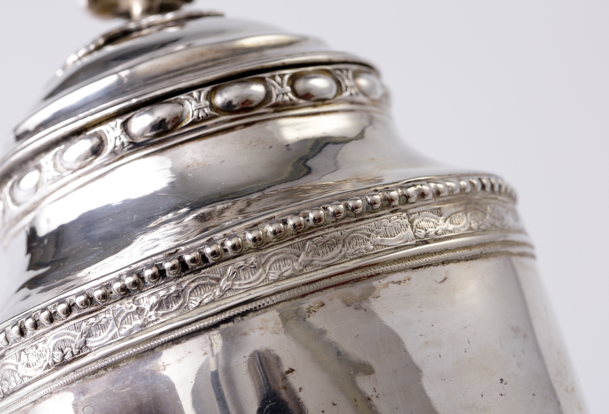 Kaffekanna i silver. 
Cylindrisk modell på tre tassfötter. Graverad bård nedtill, följd av pärlbård. Välvt lock med graverad bård, pärlbård, äggstavsbård och knopp i form av en ros. Försedd med svarvat, svärtat trähandtag, med benknopp. 
Stämplar på undersidan av kannan.