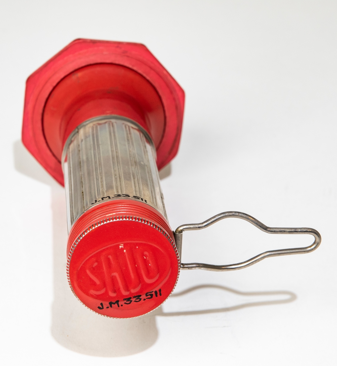 Stavlampa av plåt, gummi och glas med cylinderformad hylsa för batterier, dekorerad med räfflor. Rödlackerat skruvlock med text i relief: "SAJO". Rödlackerd, vridbar linshållare med cirkelrund glasskiva. En röd gummiring håller fast glasskivan. Hylsan är försedd med kontakter för blinkljus och stadigt ljus samt hängare av metalltråd.