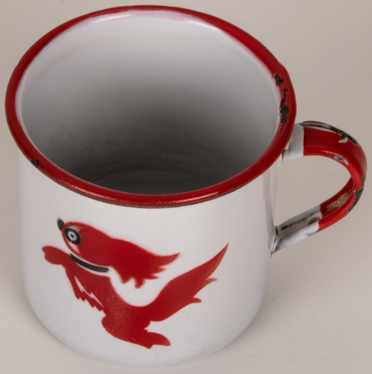 Mugg i vit emaljerad plåt. Cylindrisk med omvikt mynningskant. Lodgrepe. Kant och grepe röda. Dekor i rött och svart: hund (formgiven av Einar Nerman). Blå stämpel under: KOCKUMS (logotyp med texten K.E.R. SWEDEN) 7 cm NERMAN.