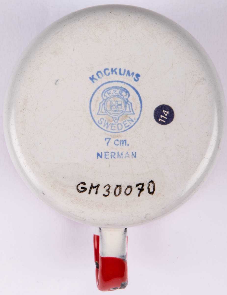 Mugg i vit emaljerad plåt. Cylindrisk med omvikt mynningskant. Lodgrepe. Kant och grepe röda. Dekor i rött och svart: hund (formgiven av Einar Nerman). Blå stämpel under: KOCKUMS (logotyp med texten K.E.R. SWEDEN) 7 cm NERMAN.