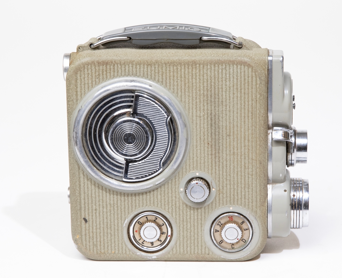 Filmkamera, dubbel-8 Eumig C 3, med kamerahus av grå metall, försedd med fjädermotor. Handtag av plast. Filmkameran är placerad i en kameraväska av brunt läder, JM 56584, och skruvas fast i väskan med en skruv. I kameraväskan förvaras ett litet linsskydd i en röd påse, och en fjärrutlösare.