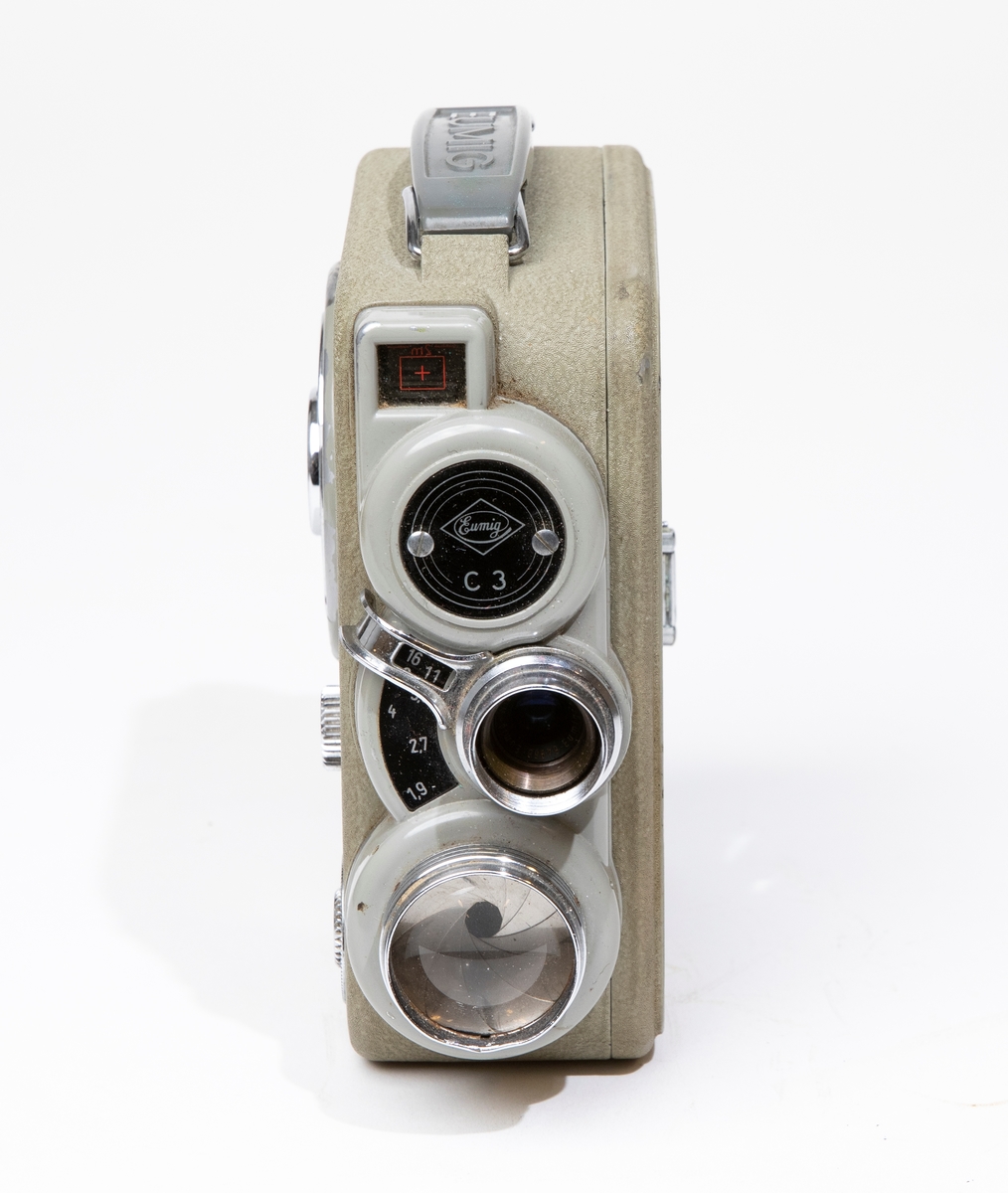 Filmkamera, dubbel-8 Eumig C 3, med kamerahus av grå metall, försedd med fjädermotor. Handtag av plast. Filmkameran är placerad i en kameraväska av brunt läder, JM 56584, och skruvas fast i väskan med en skruv. I kameraväskan förvaras ett litet linsskydd i en röd påse, och en fjärrutlösare.