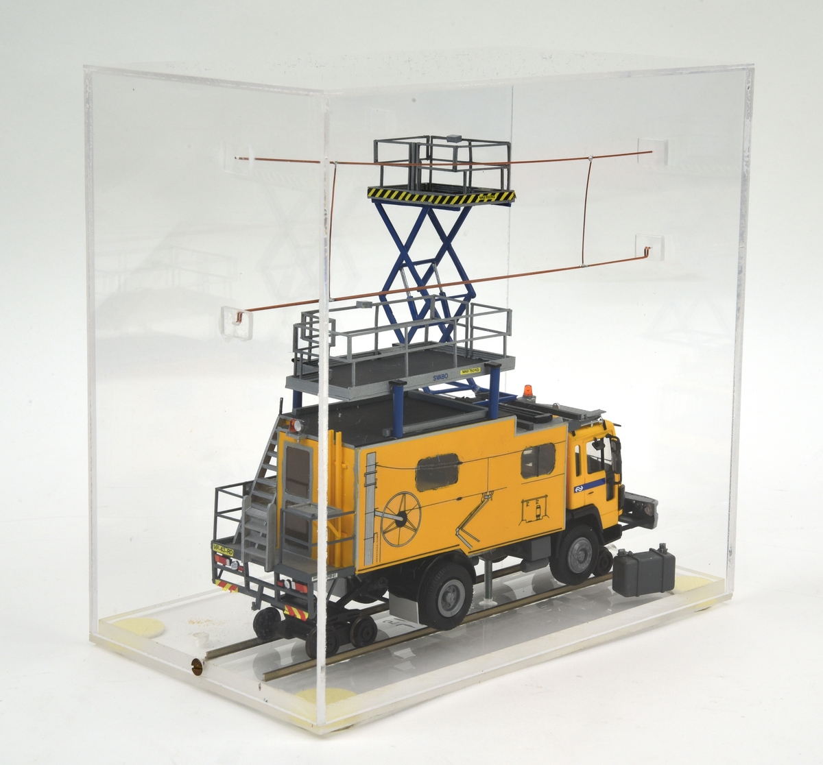 Modell i skala 1:50 av elrevisionsbil typ VRB från SRS Swedish Rail System AB baserad på ett lastbilschassi av typ Volvo FL6 för Nederlandse Spoorwegen (NS). Fordonet är gulmålat med blå detaljer i enlighet med NS:s färgsättning och på förarhyttens sidor bakom dörrarna finns NS logotyp. Flakdelen utgörs av ett skåp med fönster på sidorna samt ingång från bakre gaveln. Skåpet är gult med avbildningar av kontaktledningar. Längst bak och på skåpets tak finns fasta arbetsplattformar med skyddsräcken. På främre delan av skåpets tak finns en saxlyft med blå ben i uppfällt läge. Bilen har utöver de vanliga lastbilshjulen en enaxlad rälsboggi fram och en tvåaxlad boggi bak och modellen visar bilen i rälskörningsläge. Den främre kofångaren är framdragen och fungerar som fäste för boggien. Modellen har holländska registreringsskyltar.

Modellen är monterad i en plexiglasmonter och står på en räls av metall som är lagd direkt på monterns golv utan slipers. Ovanför bilen finns en kontakledning av koppar fäst i monterns väggar. Lastbilens bränsletank har lossnat och ligger lös i montern.