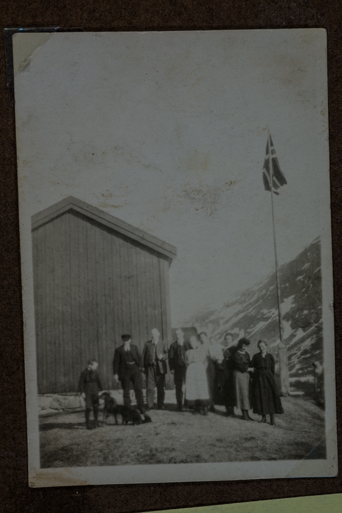 Leirfjord, Leira, Sommerseth.  På gården, 63/1, Sommerseth. Folkegruppe foran flaggstanga ved gavelveggen til bolighuset. En gutt står med to hunder. De ukjente personene er fint pyntet. Kanskje er det 17. mai.