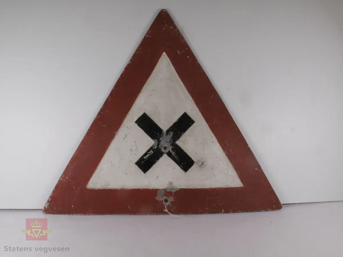 Trekantet skilt (fareskilt) med rød bord (7 cm) og svart kryss i midten. Skiltet markerer et farlig vegkryss. Skiltet bærer preg av tidens tann; flere steder har malingen skallet av. Skiltet har også fått en ruglete overflate. Det er et skruehull i midten av krysset, og to i ulike størrelser på midten nederst.