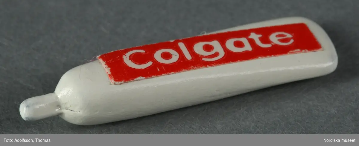 Två tandkrämstuber av märket Colgate. Den ena a) är av vitmålad avrundad metall med röd etikett och den andra b) föreställer en pappkartong och är förmodligen tillverkad av trä och papper.