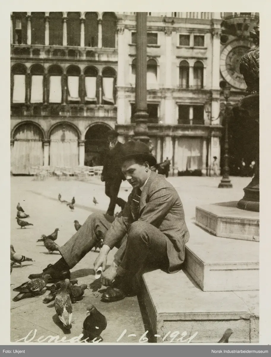 Markusplassen, Piazza San Marco. Til høyre sees halvdelen av urskiva til klokketårnet. Mann sitter på et trappetrinn med duer på åpen plass i Venezia