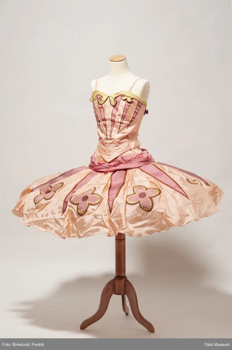Todelt ballettkjole som holdes sammen med knapper.
A Innsvingt liv, holdes sammen med hekter i ryggen. Tynne skulderstropper i strikk. Blek rosa/ ferskenfarget bunn med innfellinger i mørkere rosa. Utringing er dekorert med grønn kant. 
B: Strutteskjørt i blek rosa, fersken med innfellinger i mørkere rosa. Dekorert med applikerte blomster i rosa, gul og brun. Mørk rosa bånd i livet som knytes i sløyfe på ryggen. Skjørtet lukkes med hekter og knapper i rygg. 
C: hodepynt, avstivet, kledd i samme tekstil som resten av kostymet. Dekorert med rosa paljetter. 
D: ferskenfarget erme med innfellinger i mørkere rosa. 
E: ferskenfarget erme med innfellinger i mørkere rosa.