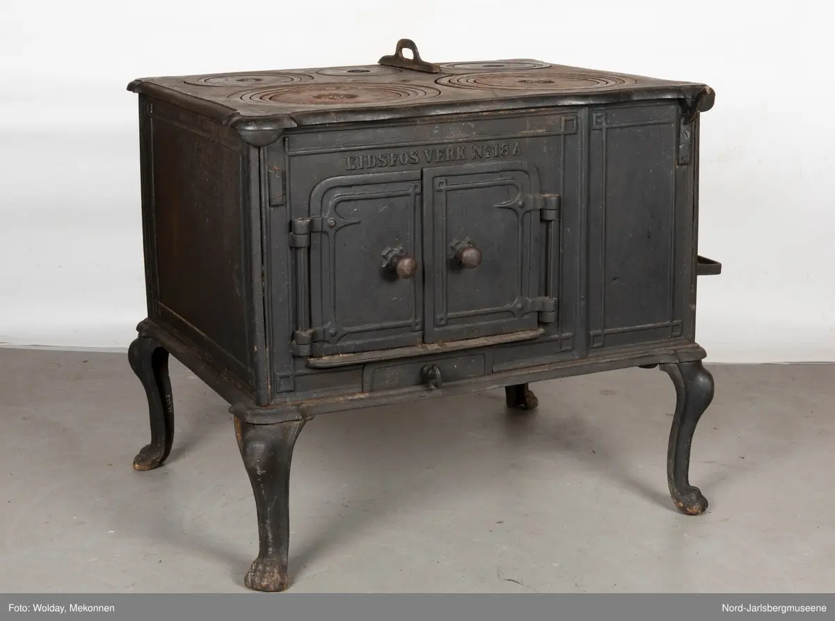 Stor, firkantet komfyr med dobbel ovnsdør til stekeovn? med støpte knotter. Enkel dekor (stilisert art nouveau). Sidedør til stekeovn. Fem kokeplater.  Bena svungne.