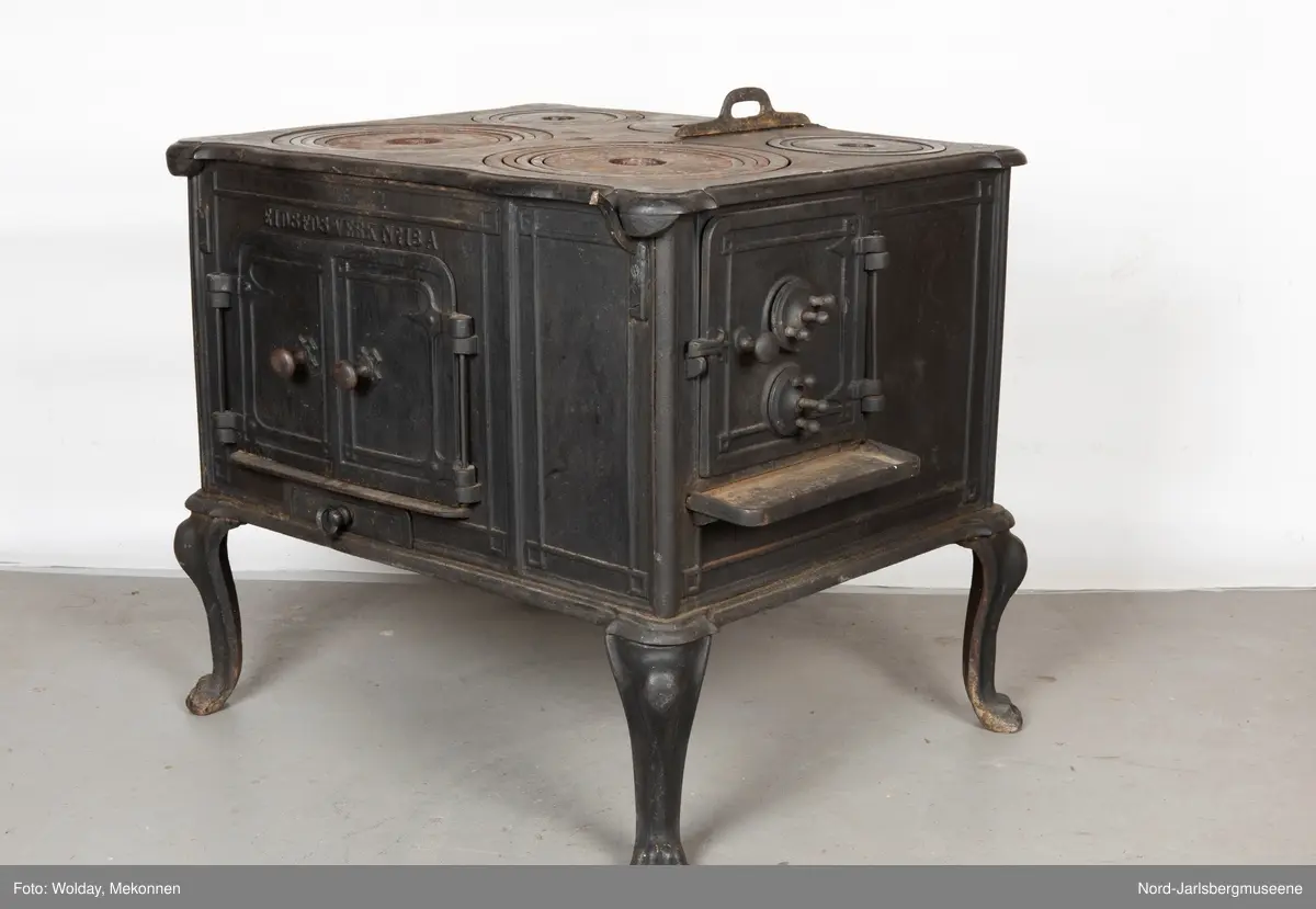 Stor, firkantet komfyr med dobbel ovnsdør til stekeovn? med støpte knotter. Enkel dekor (stilisert art nouveau). Sidedør til stekeovn. Fem kokeplater.  Bena svungne.