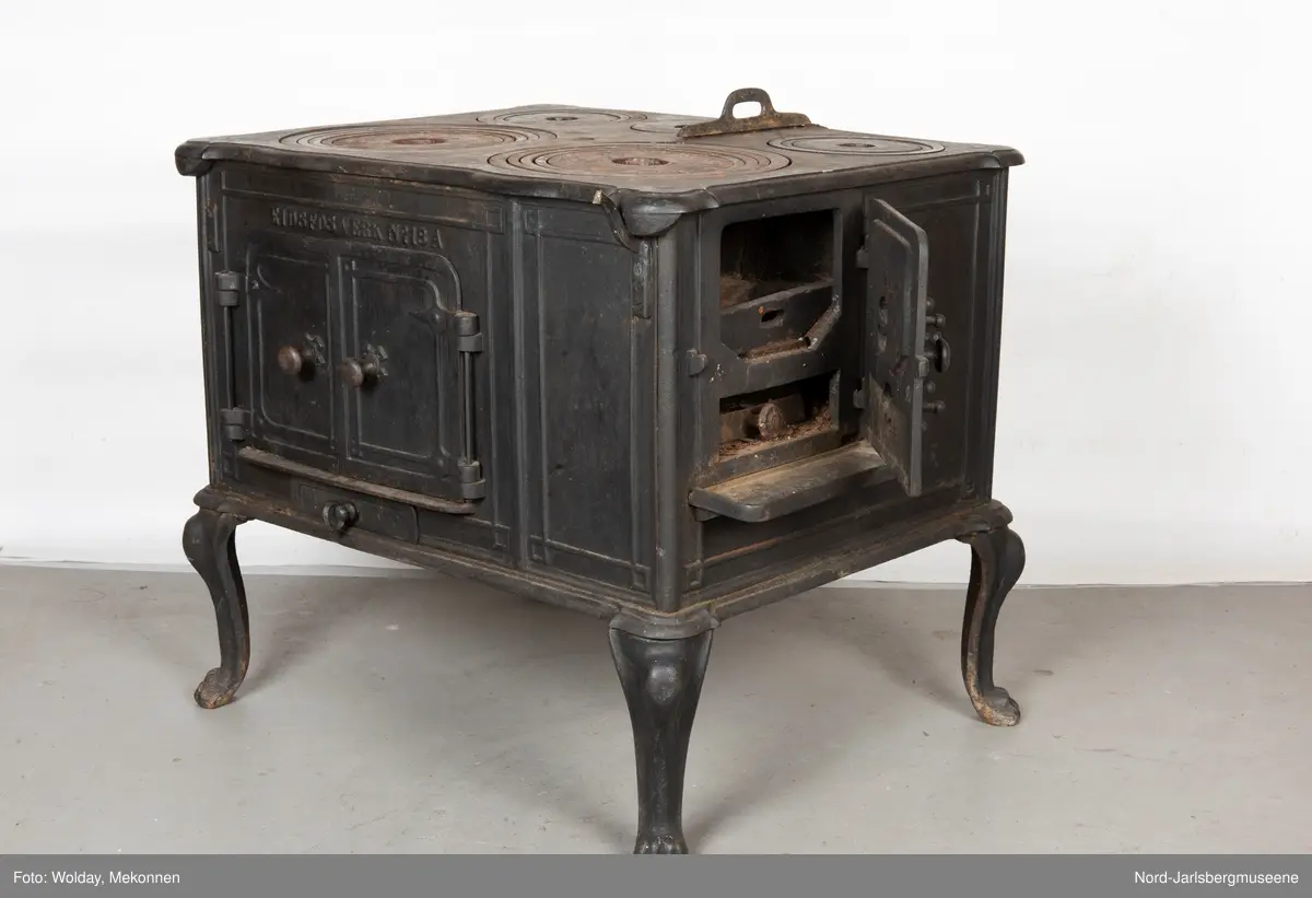Stor, firkantet komfyr med dobbel ovnsdør til stekeovn? med støpte knotter. Enkel dekor (stilisert art nouveau). Sidedør til stekeovn. Fem kokeplater.  Bena svungne.