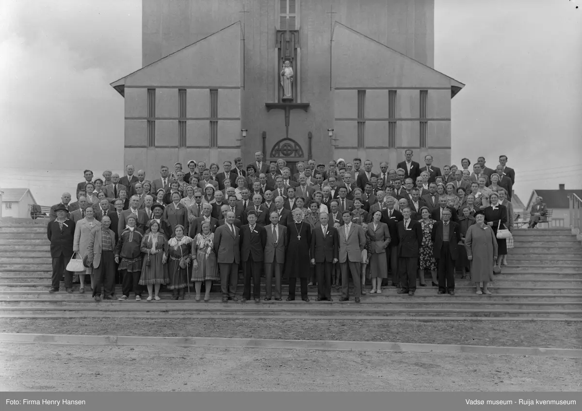 Deltakere på bispedømmemøte foran Vadsø kirke 13.8.1960. Kirka ble vigslet i 1958. Foran ser vi biskop i Finnmark Alf Wiig med iskopens surout (biskopens livkjole).  Schutte Blix (Nesseby) er han som rager høyest i bakerste rekke. ( til venstre for dørkarmen). Asbjørn Flokkmann til venstre for han. Til høyre for Blix kan det være fru Wiig, også søster Ruth Johanessen, Vadsøs helsesøster i mange år. Agnes Øwre bak henne med kofte og stokk.  Sogneprest Arvid Nergård (Tana) står to rader foran henne som har hvit hatt i bakrekka (tre-fire plasser til høyre for Hildonen)