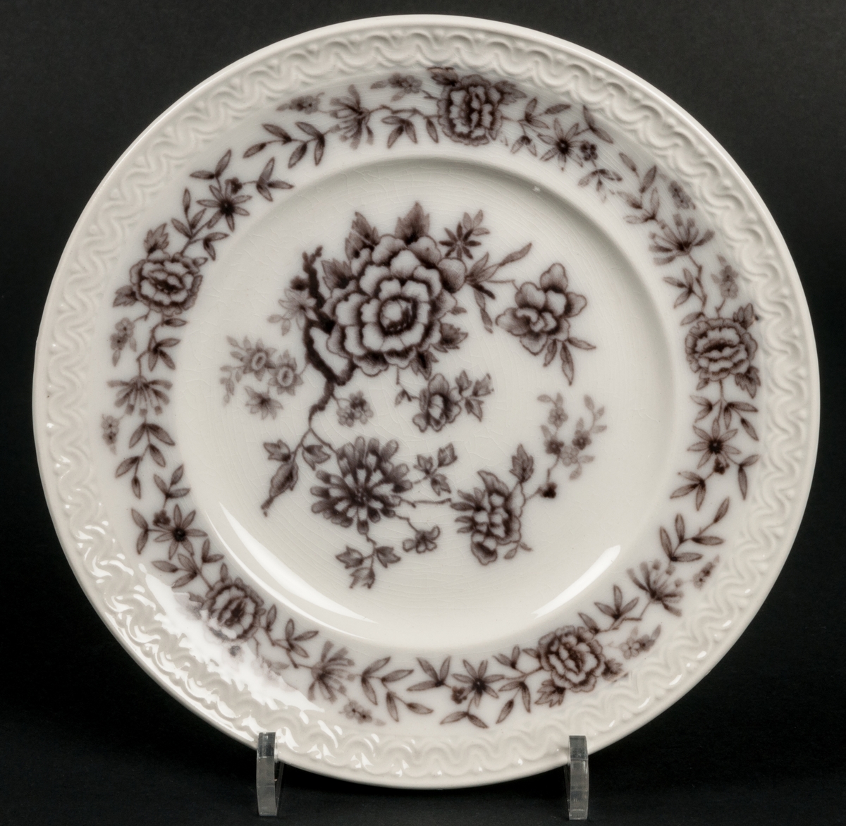 Assiett dekor Stjärnsund. Koppargravyrtryck i mullbärsbrunt.
Vit massa med brun dekor av blomster.
Bård i godset längs brämet.