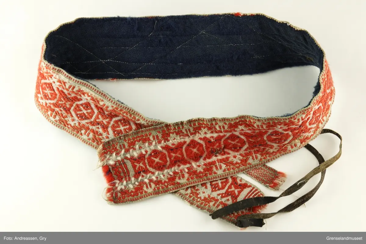 Vevd på grindvev i tre deler som er sydd sammen på langs. Sidestykkene har likt mønster. Bunn i hvitt bomullsgarn, dekor i rødt og litt blått plukk. Renningsrips. Lukning består av skinnhempe i den ene enden og skinnsnor i den andre enden. Beltet er foret med blått ullstoff.