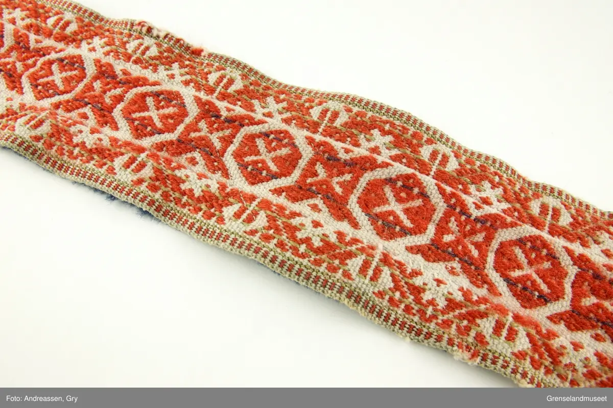 Vevd på grindvev i tre deler som er sydd sammen på langs. Sidestykkene har likt mønster. Bunn i hvitt bomullsgarn, dekor i rødt og litt blått plukk. Renningsrips. Lukning består av skinnhempe i den ene enden og skinnsnor i den andre enden. Beltet er foret med blått ullstoff.