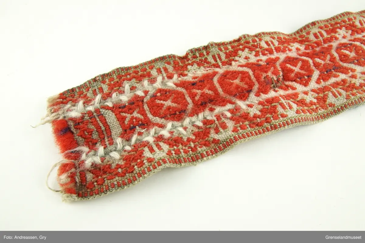 Vevd på grindvev i tre deler som er sydd sammen på langs. Sidestykkene har likt mønster. Bunn i hvitt bomullsgarn, dekor i rødt og litt blått plukk. Renningsrips. Lukning består av skinnhempe i den ene enden og skinnsnor i den andre enden. Beltet er foret med blått ullstoff.