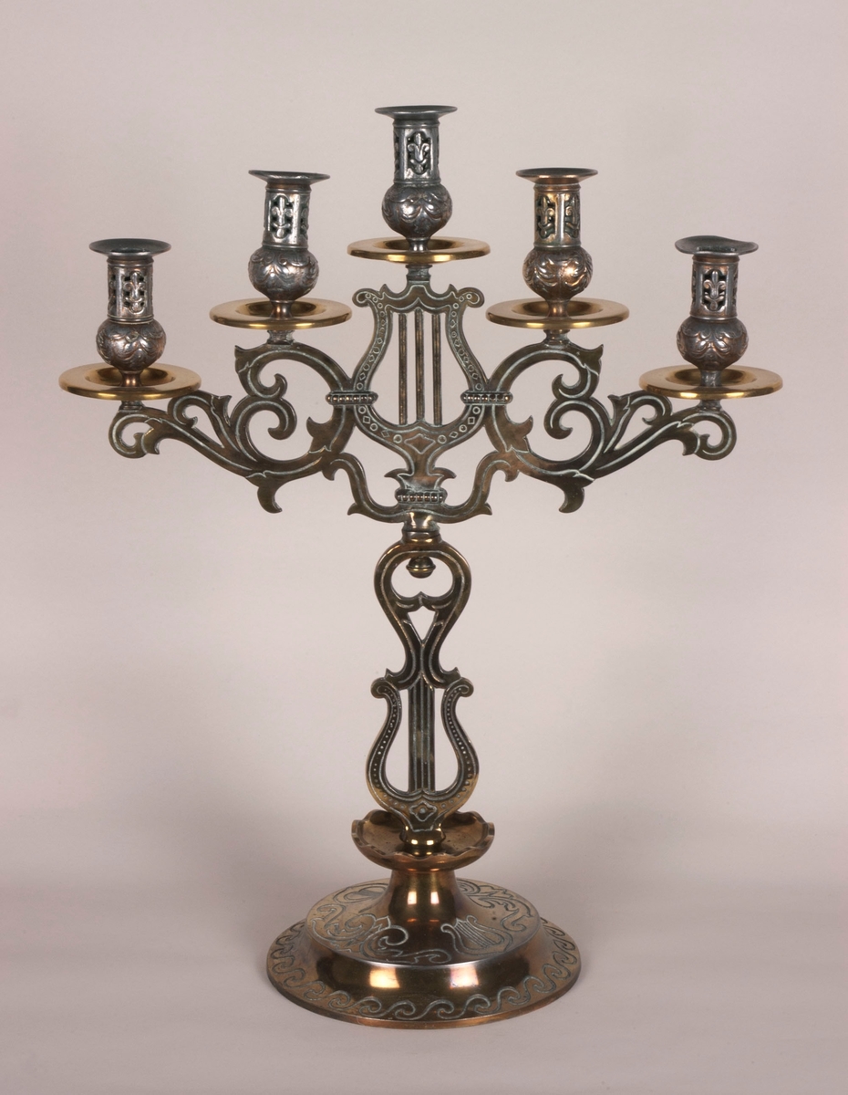 Femarmet lysestake. Rund sokkel. Fem armer springer ut fra dem midtre stolpen, som består av to lyrer. Armene og stolpen er av støpt messing. Lysholderne er i annet materiale, muligens nikkel. Dekorasjoner i hele stolpen, armene og i alle lysholderne.