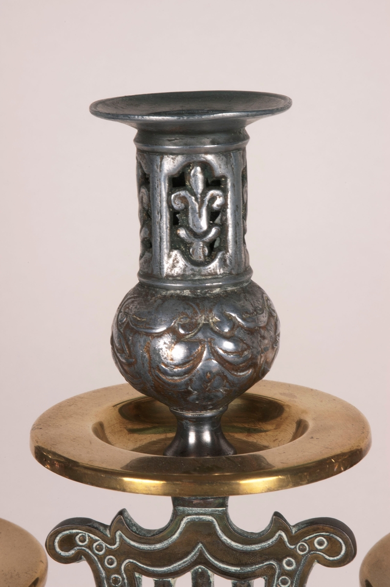 Femarmet lysestake. Rund sokkel. Fem armer springer ut fra dem midtre stolpen, som består av to lyrer. Armene og stolpen er av støpt messing. Lysholderne er i annet materiale, muligens nikkel. Dekorasjoner i hele stolpen, armene og i alle lysholderne.