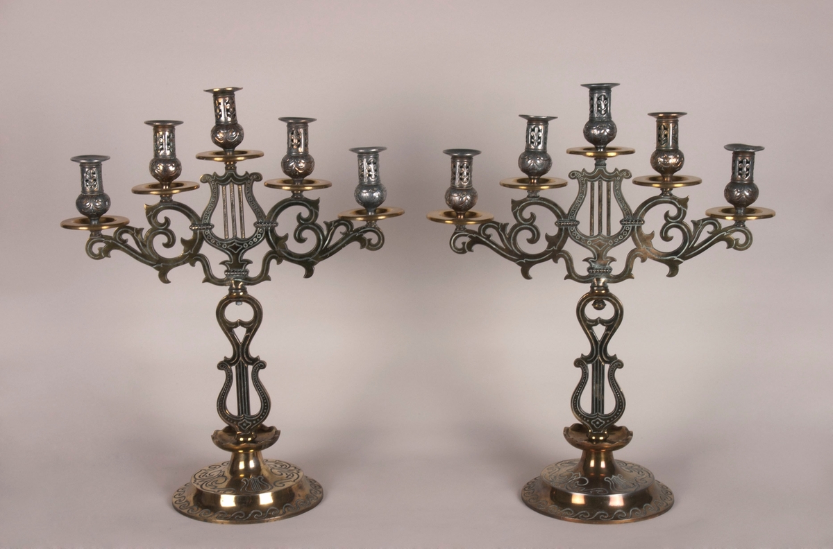 Femarmet lysestake. Rund sokkel. Fem armer springer ut fra dem midtre stolpen, som består av to lyrer. Armene og stolpen er av støpt messing. Lysholderne er i annet materiale, muligens nikkel. Dekorasjoner i hele stolpen, armene og i alle lysholderne.