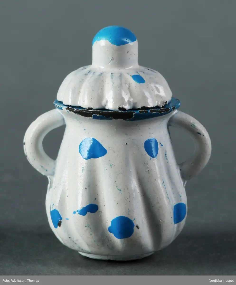 Vit kaffeservis med turkos dekor i form av prickar: a) kaffekanna, b) gräddkanna, c:1-2) sockerskål med lock, d-g) fyra koppar och h-k) fyra fat. Tillverkade av metall. Kaffeservisen ska stå på matbordet (NM.0333310 a) mitt i matsalen i dockskåpet NM.0331721+.