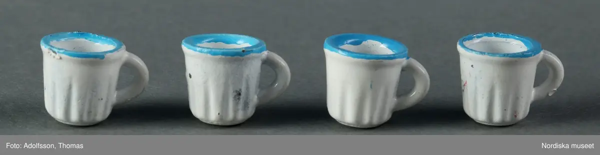 Vit kaffeservis med turkos dekor i form av prickar: a) kaffekanna, b) gräddkanna, c:1-2) sockerskål med lock, d-g) fyra koppar och h-k) fyra fat. Tillverkade av metall. Kaffeservisen ska stå på matbordet (NM.0333310 a) mitt i matsalen i dockskåpet NM.0331721+.