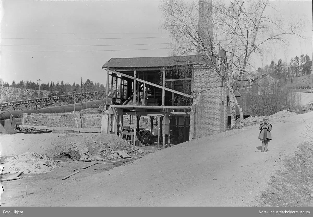 Anleggsarbeid under bygging av Labro Kraftstasjon. Bygg med høy murpipe under oppføring. Barn oppstilt på veien ved byggeplassen.