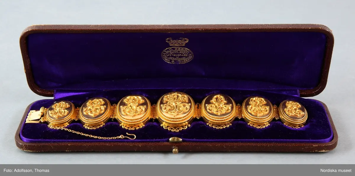 A) Armband, av matt och blankt guld, bestående av sju stående, sammanlänkade medaljonger som innesluter fotografiporträtt under glas. På varje medaljonglock ett pålagt monogram (för- och efternamnsinitialer) samt runt ytterkanten en smal pärlstav.  Genombruten dekor upptill och nedtill. Släta baksidor. Lås med bred snäppare stämplad med firmanamn, riks-, karat-, och stadsstämpel samt årsbokstav. Säkerhetskedja, med hake. Tillverkat i Stockholm 1875 av L C Féron/firma Gustaf Möllenborg. 

B) Originaletui, avlångt, välvt, med rundade hörn, klätt med brunt mönsterpressat skinn. Fällock med tryckknäppe av mässing. Etuiets insida klädd med lila sammet och siden (locket). Firmastämpel i guldtryck.

Porträtten föreställer Victor Edman (4) i centrum, omgiven av sina sex barn. På vänster sida finns de två äldsta barnen (2) och (3) från hans första äktenskap med Ulrika Charlotta Edman, f. Lagerhjelm (1818-1846). Längst ut på vänster sida finns det första barnet (1) i hans andra äktenskap med Emilia Elisabeth (Elise) Edman, f. Biel, en son som dog i unga år. På höger sida, de yngre barnen (5) (6) och (7), med Elise Edman:
1. VE: Johan Victor Edman, student, f. 12/8 1852, d. 6/3 1866.
2. CE: Carl Gustaf Edman, kanslisekreterare i civildepartementet, f. 26/7 1846, d. 3/5 1921. 
3. PE: Pehr Victor Edman, kapten vid Västmanlands regemente, f. 10/10 1841, d. 7/12 1879.
4. VE: Beställaren av armbandet, familjefadern Victor Edman, överstelöjtnant, f. 29/9 1813, d. 6/10 1880.  I locket en ljus lock av Victor Edmans hår. 
5. AE: Anna Mathilda Elisabeth Edman, f. 4/6 1856, d. 13/9 1882. I locket ett porträtt av maken, Anders Axel Altberg, godsägare (1839-1902). 
6. GE: Fredrik Gillis Edman, agronom, f. 6/7 1857, d. 19/11 1902.
7. EE: Hedvig Emilia (Emy) Edman, f. 2/5 1865, d. 11/9 1935, senare trägravör. Gift 1892 med Johan (John) Ludvig Kindborg, konstnär (1861-1907). 
/Anna Womack 2017-08-18