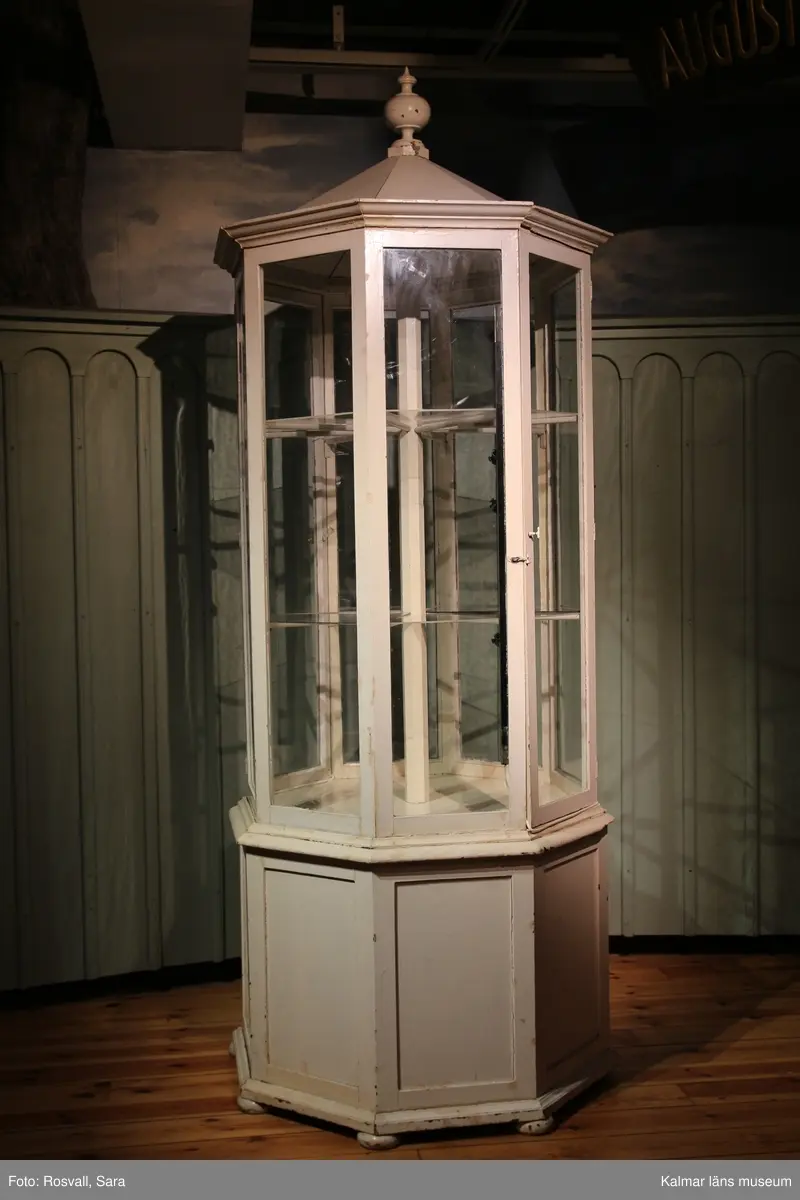 KLM 28082:491. Monter, trä, glas, metall. Åttakantig vitmålad golvmonter med täckt ihåligt underrede av trä med en dörr. Åtta glasrutor varara två med dörrar. Två nivåer med hyllplan, bestående av lösa trekantiga glashyllor på varje plan. Täckt överdel av trä med knopp.