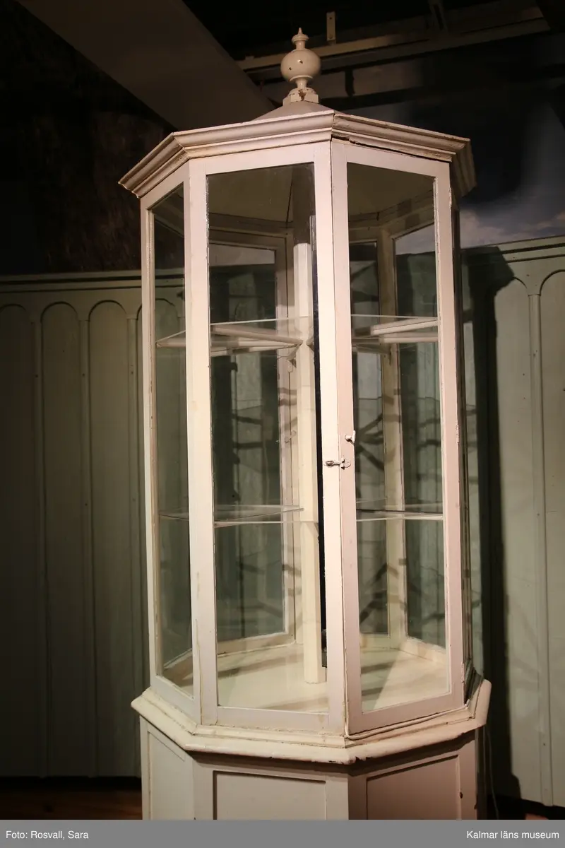 KLM 28082:491. Monter, trä, glas, metall. Åttakantig vitmålad golvmonter med täckt ihåligt underrede av trä med en dörr. Åtta glasrutor varara två med dörrar. Två nivåer med hyllplan, bestående av lösa trekantiga glashyllor på varje plan. Täckt överdel av trä med knopp.