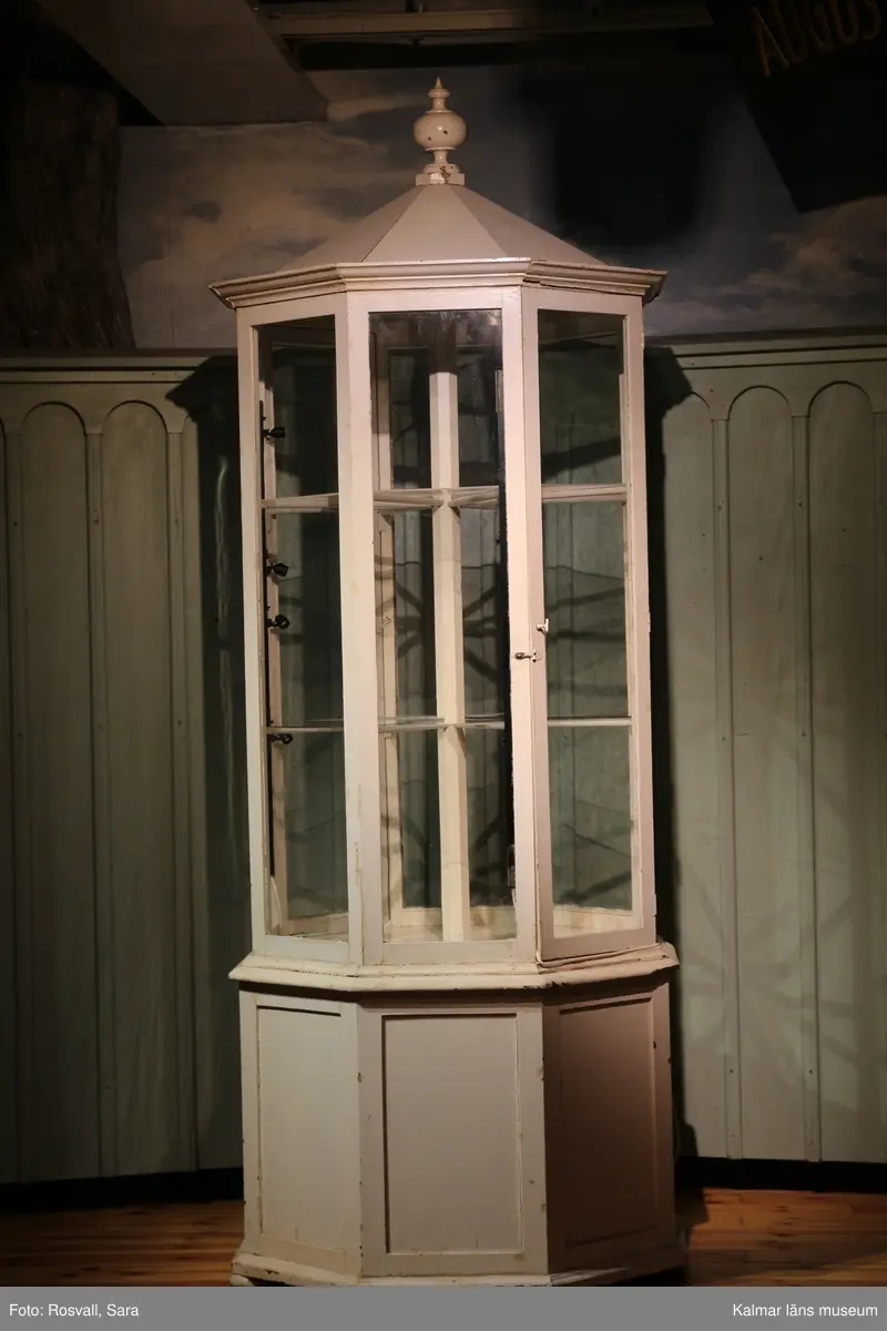 KLM 28082:491. Monter, trä, glas, metall. Åttakantig vitmålad golvmonter med täckt ihåligt underrede av trä med en dörr. Åtta glasrutor varara två med dörrar. Två nivåer med hyllplan, bestående av lösa trekantiga glashyllor på varje plan. Täckt överdel av trä med knopp.