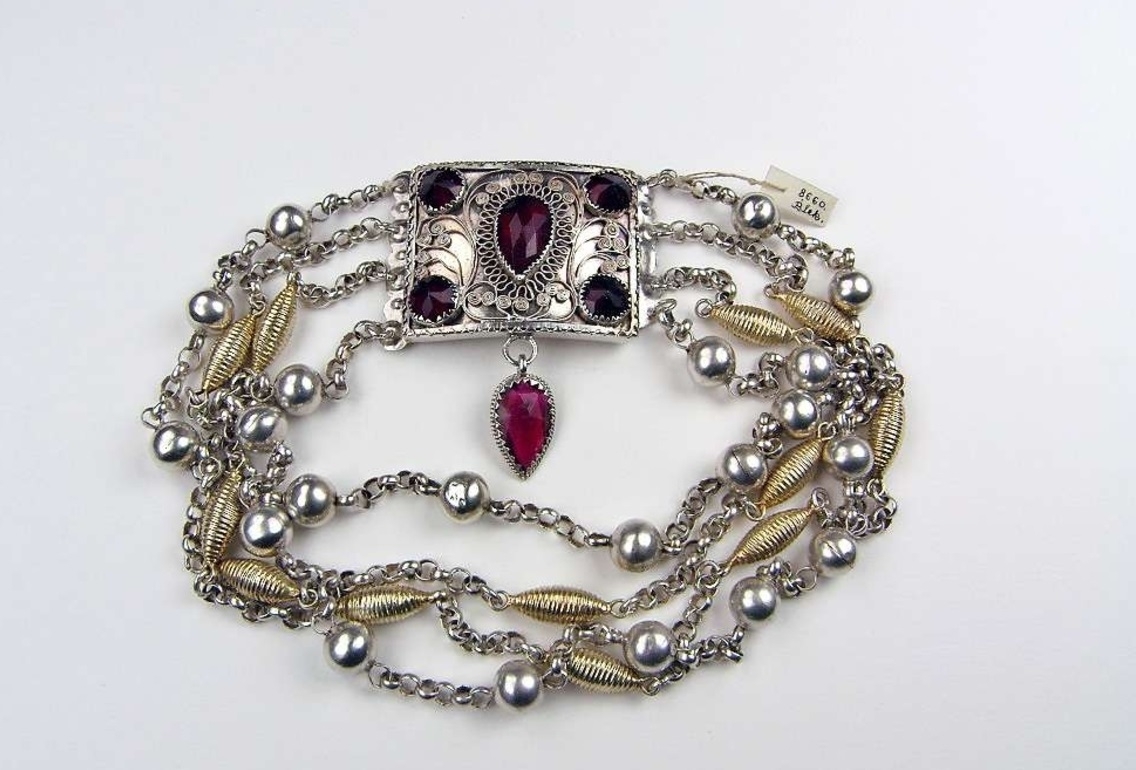 Halslås, av silver, rektangulärt med instickslås.  På låsets ovansida gles filigrandekor, i varje hörna en rund, fasettslipad, röd glassten i klofattning, mitt på en droppformad dito. Från låsets nederkant, fäst i en ögla, hänger en droppformad, fasettslipad röd glassten i klofattning. Fyra rader kedjor, ankarlänk, de yttre med runda silverkulor placerade med ca. tre cm mellanrum, de två inre med förgyllda spolar som dekor. Under bottenplattan svisselerat initialerna EJD. Mästarstämpel IMH, Johan Mathias Holm verksam i Karlskrona 1817-1830.