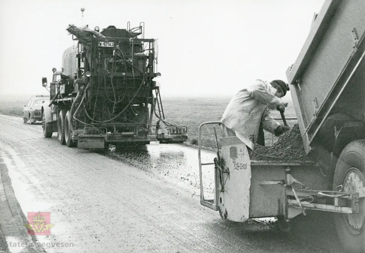 Opplæring med sprøytetank på Volvo lastebil med kjennemerke W-92563, Statens vegvesen Nordland 510-05. Testkjøring på fv på Fauskemyra 1977. En Ford Transit står bak en 1976 modell Audi 80 personbil. Mercedes-Benz lastebil foran veivalsen. Mannskapet i skyggelue og arbeidstøy (overaller) står ved bilene. 

Bilde 2) I arbeid på vegen. Mannen på sprøytebilen er godt tildekket. NVM 18-F-00414-1