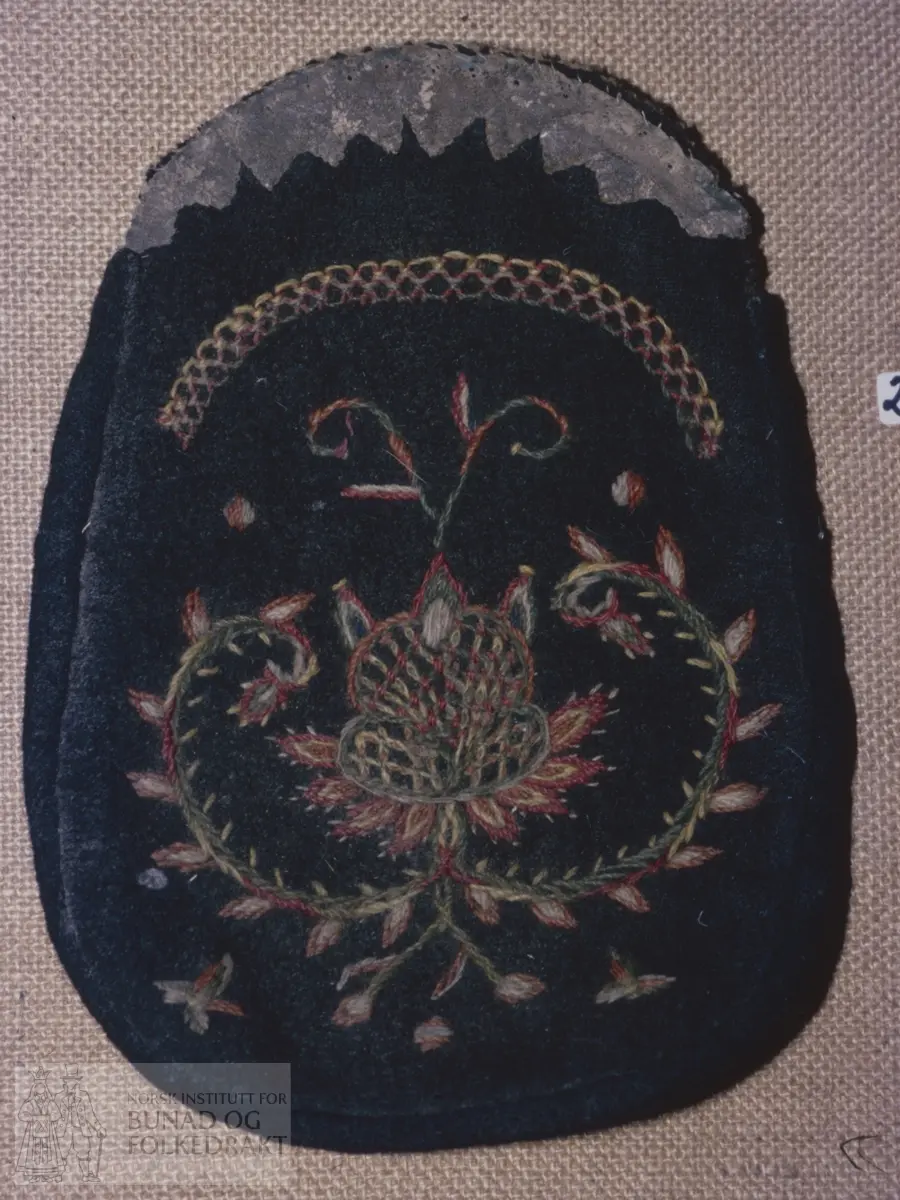 Mørkebrun vadmål på framsida pryda med rosemotiv og sauma med tynt, totråds ullgarn i raudt, grønt, gult og grått. Kontursting. "Taggekant" i skinn øvst. Skinn også på baksida. Fôra med grovt linlerret. Sauma med ubleika lingarn. Høyde:  25 cm.