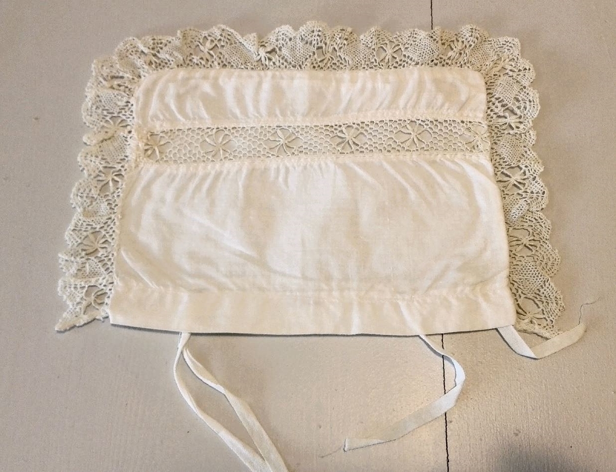 Sydd av kvitt bomullslereft med mellomverk av 2 cm brei blonde. Kanta med 3 cm brei tunga blonde kring 3 kantar. 2 sett bendelband i åpning på eine langsida.