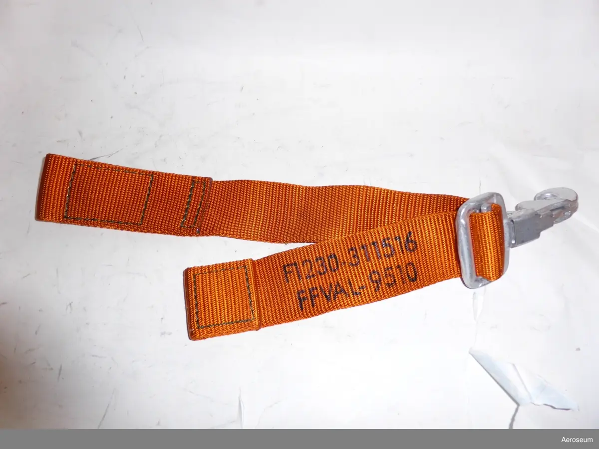 En fastbindningsrem för helikopter. är orange och i svart text står det på den: "F1230-311516 FFVAL-9510". Har även en metallhake.
