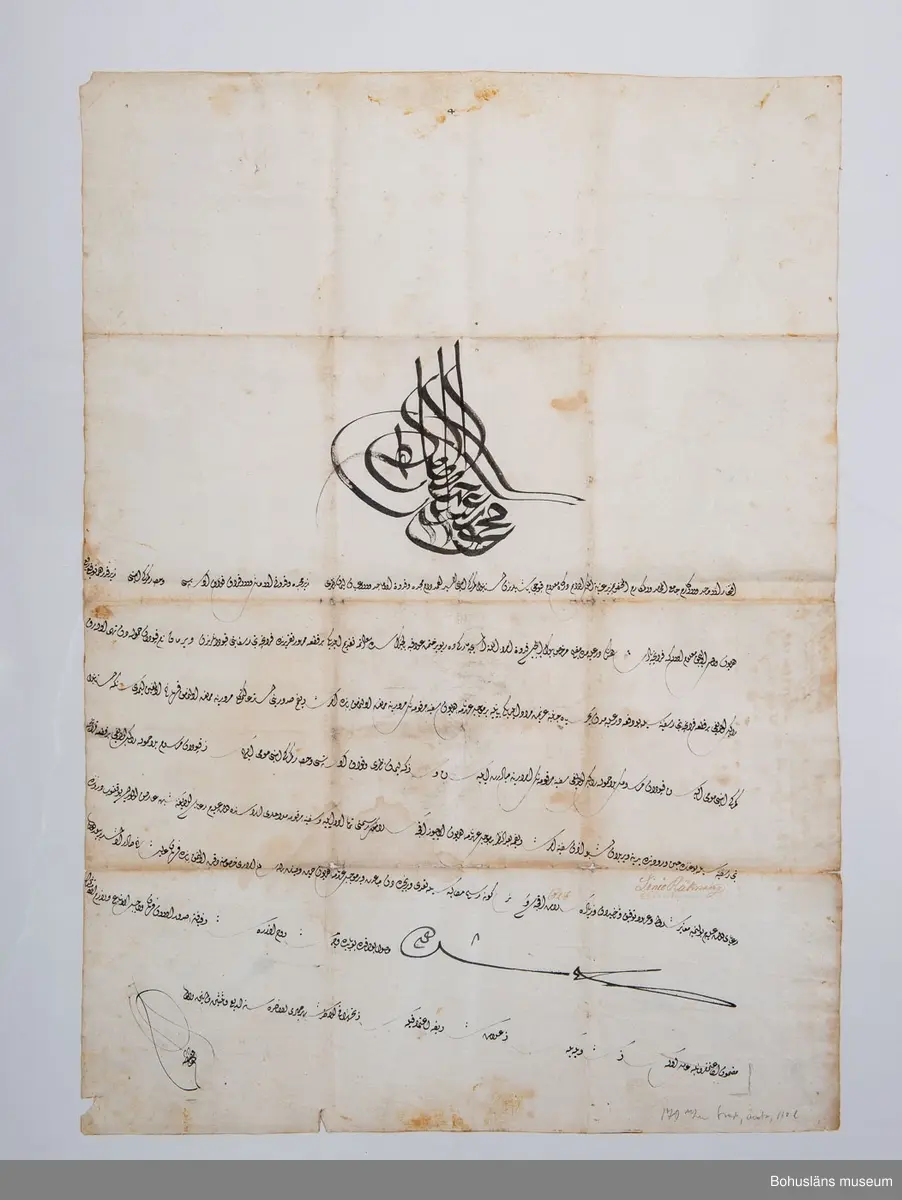 Kraftigt vitt pappersark, svagt vaxat, något snett skuret och i 16 vikningar.

Uppgifter på framsidan/insidan:
Text på arabiska skrivet med svart bläck täckande 3/4 av arket i jämn och välskriven kalligrafi där varje rad avslutas snett upp åt vänster. Överst en stor centralt placerad anfang (namnschiffer) med guldglitter fästad på färgen. Åtta rader text undertecknad med två namnteckningar.

Uppgifter på baksidan på vad som har varit utsida av fermanen då den varit ihopvikt till storlek motsvarande ungefär ett A5-kuvert:
Skrivet med svart bläck: kortfattad arabisk utanpåskrift  på tre vikningar.

Skrivet med brunt bläck: 
1823 den 16 Januari
Foglekjärr Uti  Bro
nr 26 januari 
1824
1712
1451

[Flera oläsliga rader] den 7 augusti 1822
1821 [oläsligt]
1821 [oläsligt]
Åhr 1824 den den 20de Julli
Lündby Heden.  i Skånne
Län

Vikningar, missfärgning efter vikningar, fläckar.

I 1869 års tryckta katalog över Uddevalla Musei Historiska Samlingar främmande nationers skrift:
Tre turkiska firmaner för fartyg.