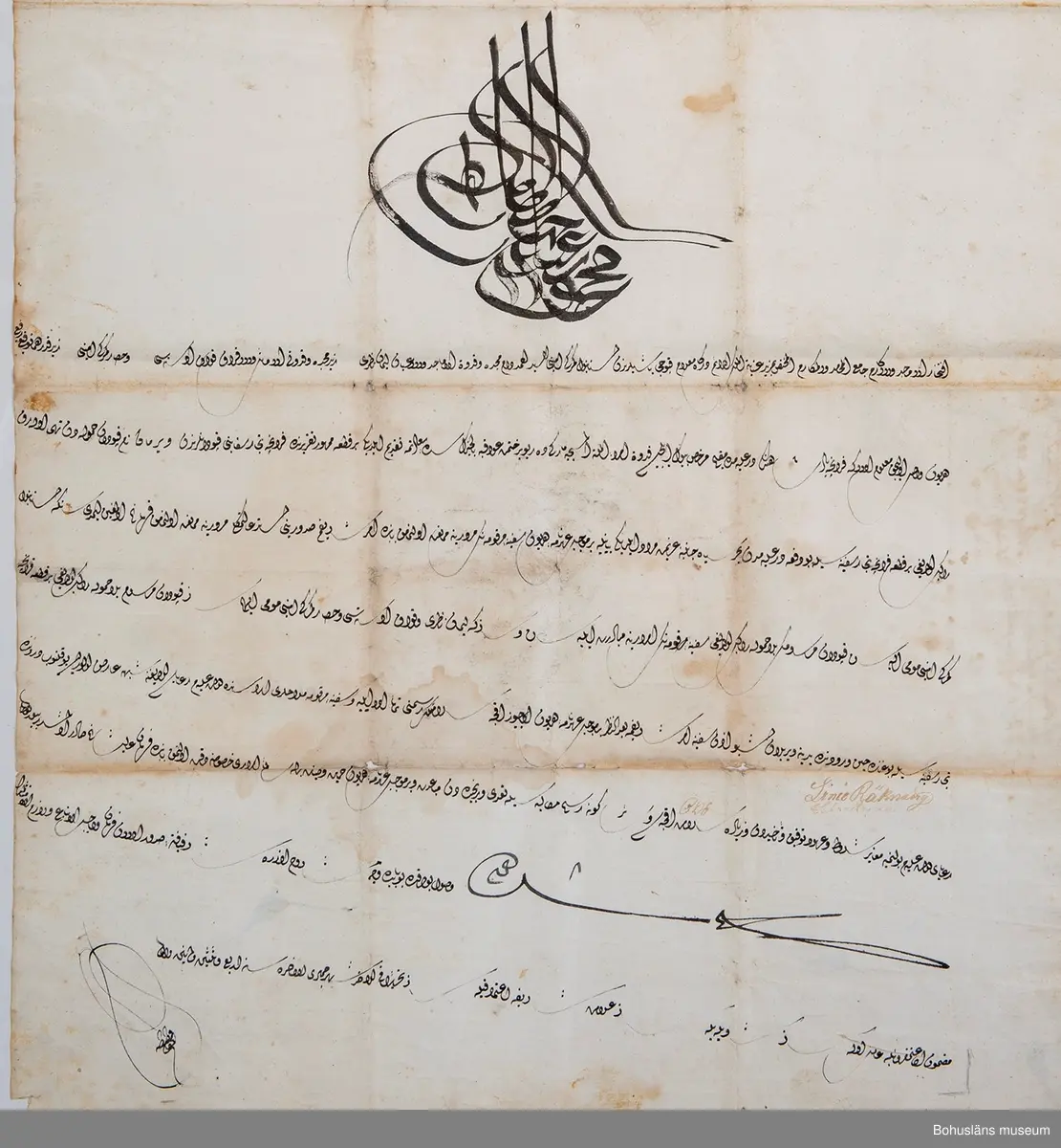 Kraftigt vitt pappersark, svagt vaxat, något snett skuret och i 16 vikningar.

Uppgifter på framsidan/insidan:
Text på arabiska skrivet med svart bläck täckande 3/4 av arket i jämn och välskriven kalligrafi där varje rad avslutas snett upp åt vänster. Överst en stor centralt placerad anfang (namnschiffer) med guldglitter fästad på färgen. Åtta rader text undertecknad med två namnteckningar.

Uppgifter på baksidan på vad som har varit utsida av fermanen då den varit ihopvikt till storlek motsvarande ungefär ett A5-kuvert:
Skrivet med svart bläck: kortfattad arabisk utanpåskrift  på tre vikningar.

Skrivet med brunt bläck: 
1823 den 16 Januari
Foglekjärr Uti  Bro
nr 26 januari 
1824
1712
1451

[Flera oläsliga rader] den 7 augusti 1822
1821 [oläsligt]
1821 [oläsligt]
Åhr 1824 den den 20de Julli
Lündby Heden.  i Skånne
Län

Vikningar, missfärgning efter vikningar, fläckar.

I 1869 års tryckta katalog över Uddevalla Musei Historiska Samlingar främmande nationers skrift:
Tre turkiska firmaner för fartyg.